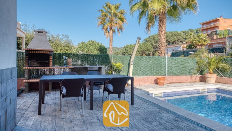 Vakantievilla's Costa Brava Spanje - Villa Zarita - Eetgedeelte buiten bij het zwembad van Villa Zarita, voorzien van een donkere Wicker tuintafel, vier stoelen, een ingebouwde stenen barbecue en palmbomen onder een blauwe lucht.