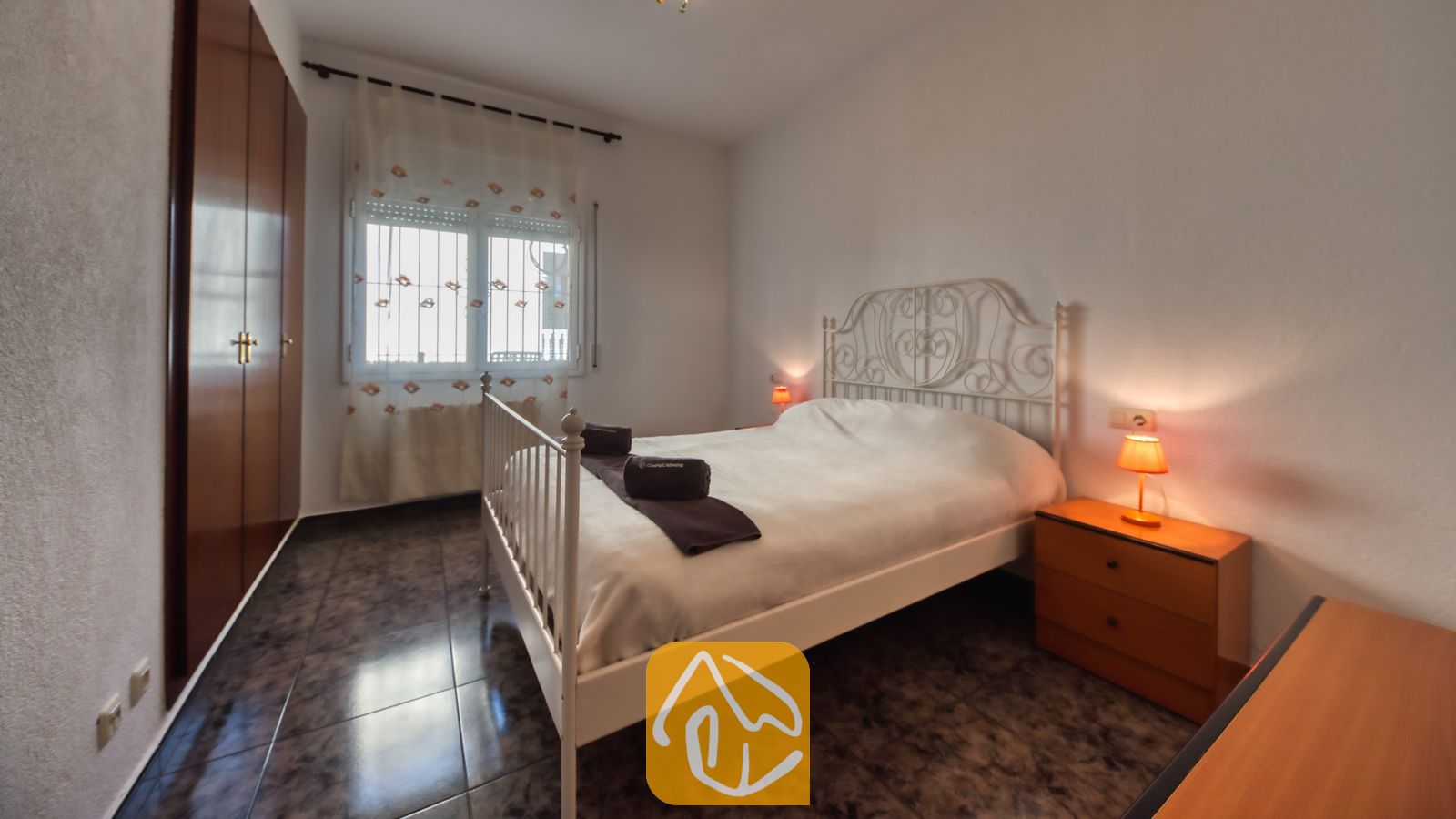 Vakantievilla's Costa Brava Spanje - Villa Zarita - Slaapkamer met een wit metalen bed, donkere tegelvloer, houten nachtkastje met een oranje lamp en een inbouwkast.