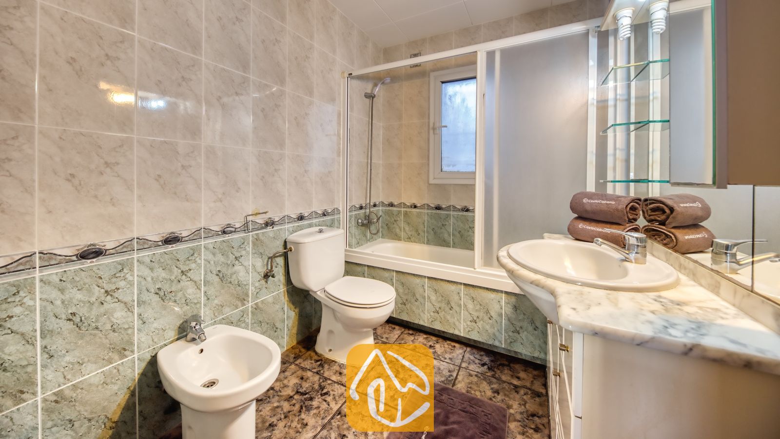 Vakantievilla's Costa Brava Spanje - Villa Zarita - Badkamer met muren van marmerlook tegels, een wit badmeubel met een waskom op het blad, een toilet, een bidet en een ligbad met glazen schuifdeuren.