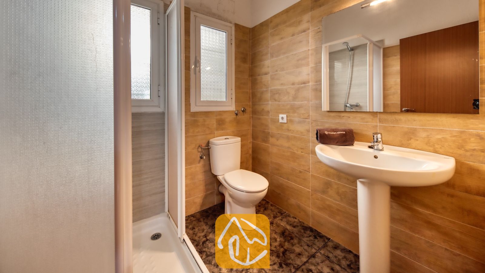 Vakantievilla's Costa Brava Spanje - Villa Zarita - Badkamer met beige betegelde muren, een witte wastafel op zuil met een bruine handdoek, een toilet en een inloopdouche met glazen deur.