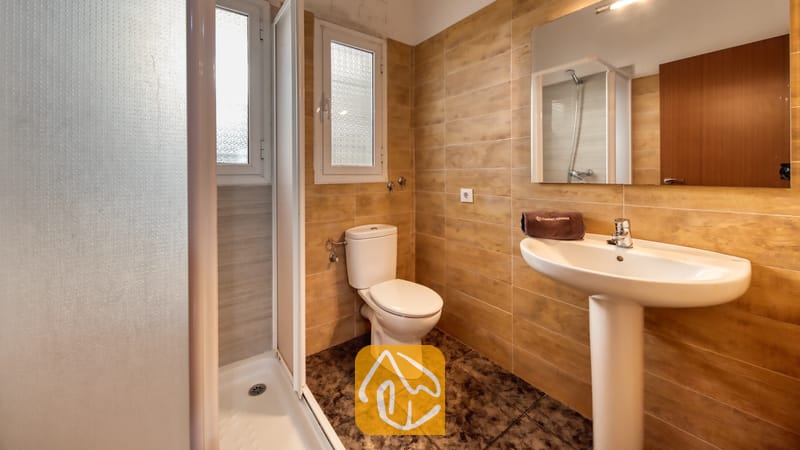 Holiday villas Costa Brava Spain - Villa Zarita - Bathroom featuring beige tiled walls, a white pedestal sink with a brown towel, a toilet, and a glass-door shower stall.
