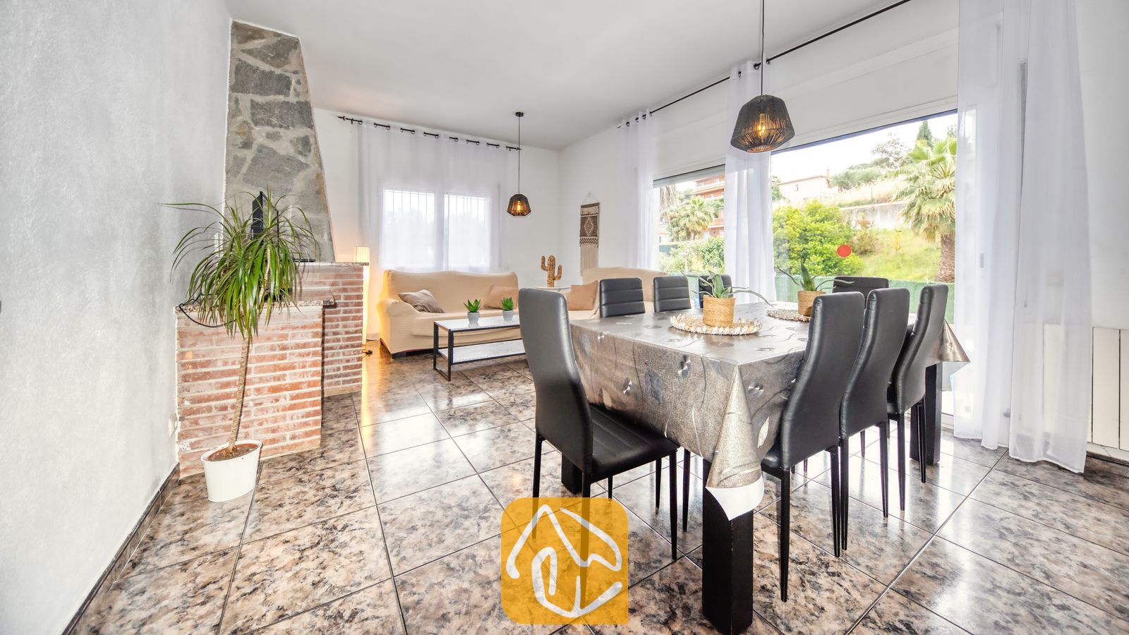 Vakantievilla's Costa Brava Spanje - Villa Zarita - Ruime woon- en eetkamer in Villa Zarita, voorzien van een stenen open haard, plavuizen vloeren, een eettafel met zwarte stoelen en grote ramen met uitzicht op de buitenlucht.