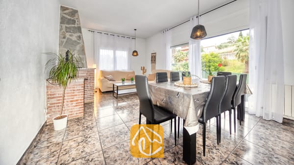 Vakantievilla's Costa Brava Spanje - Villa Zarita - Ruime woon- en eetkamer in Villa Zarita, voorzien van een stenen open haard, plavuizen vloeren, een eettafel met zwarte stoelen en grote ramen met uitzicht op de buitenlucht.