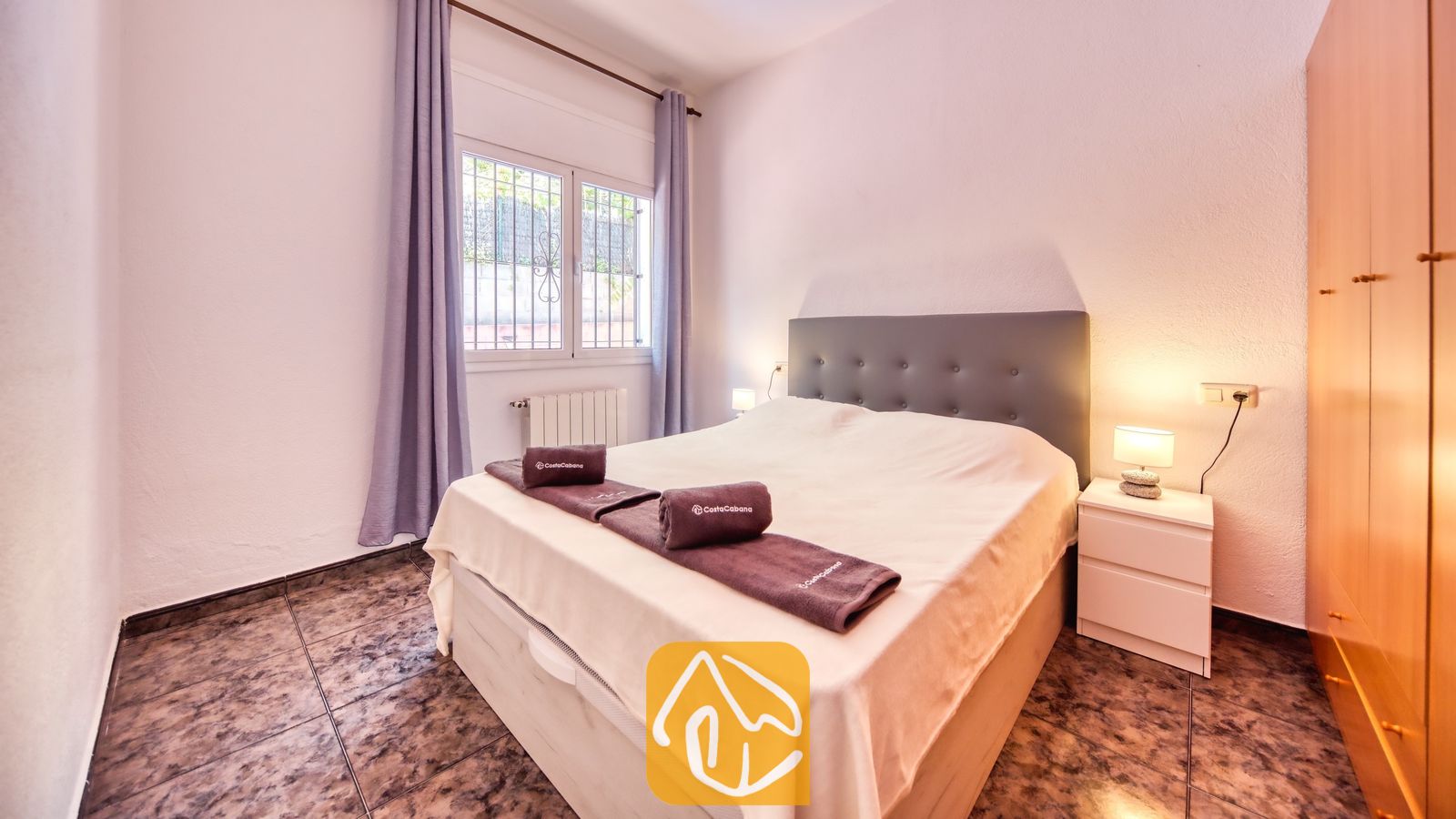 Vakantievilla's Costa Brava Spanje - Villa Zarita - Slaapkamer met een tweepersoonsbed, donkergrijze gestoffeerde hoofdbord, opgerolde handdoeken, witte nachtkastjes met lampen, paarse gordijnen en een tegelvloer.