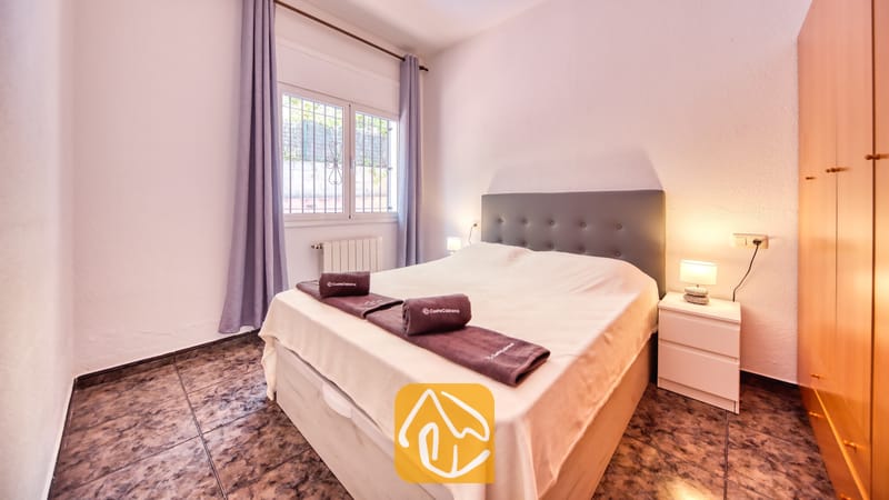 Vakantievilla's Costa Brava Spanje - Villa Zarita - Slaapkamer met een tweepersoonsbed, donkergrijze gestoffeerde hoofdbord, opgerolde handdoeken, witte nachtkastjes met lampen, paarse gordijnen en een tegelvloer.