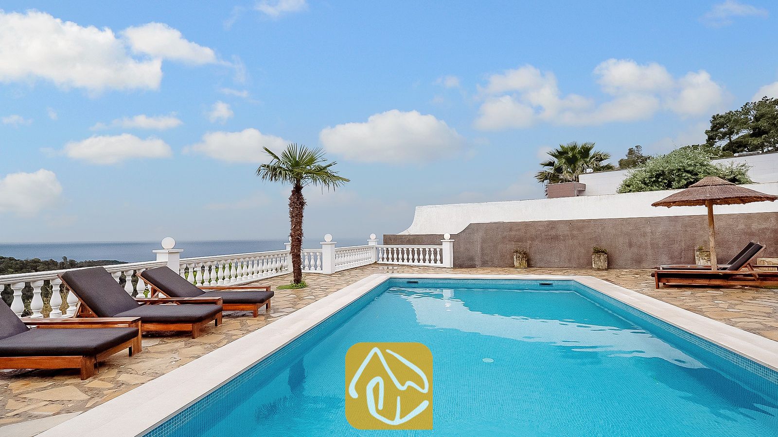Vakantievilla's Costa Brava Spanje - Villa Tropicana - Privézwembad bij Villa Tropicana, omringd door stenen tegels, houten ligstoelen met donkere kussens en een witte balustrade met uitzicht op zee onder een blauwe lucht.