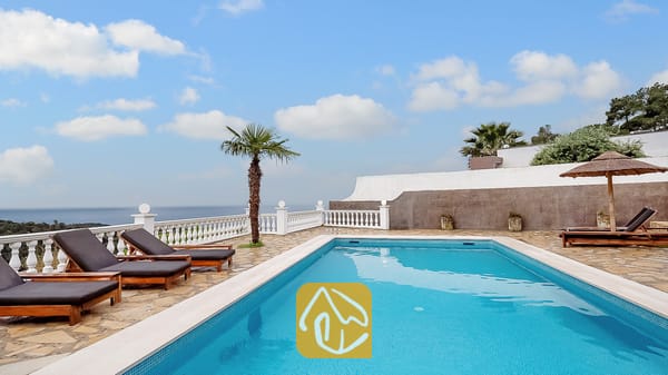 Vakantievilla's Costa Brava Spanje - Villa Tropicana - Privézwembad bij Villa Tropicana, omringd door stenen tegels, houten ligstoelen met donkere kussens en een witte balustrade met uitzicht op zee onder een blauwe lucht.