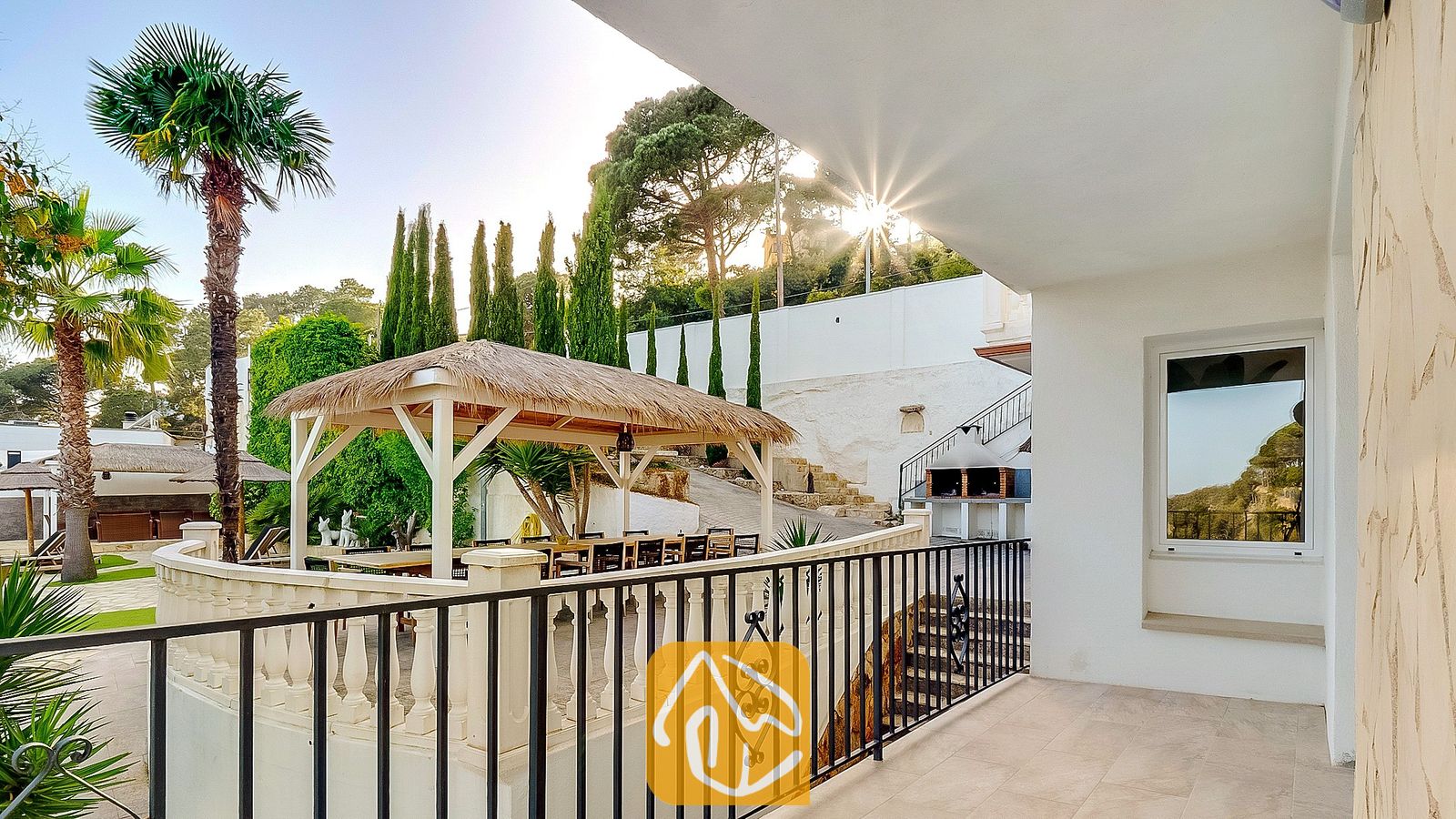 Vakantievilla's Costa Brava Spanje - Villa Tropicana - Overdekt terras bij Villa Tropicana met uitzicht op een rieten gazebo, een buiten-eethoek, palmbomen en een stenen barbecue tegen een heuvel.