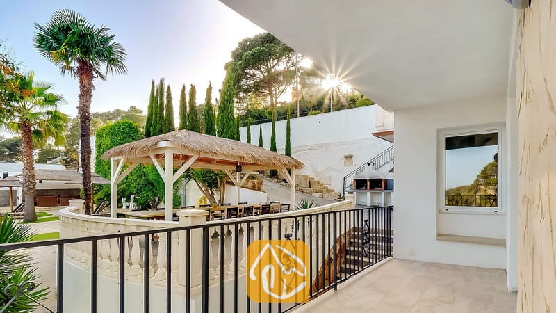 Vakantievilla's Costa Brava Spanje - Villa Tropicana - Overdekt terras bij Villa Tropicana met uitzicht op een rieten gazebo, een buiten-eethoek, palmbomen en een stenen barbecue tegen een heuvel.