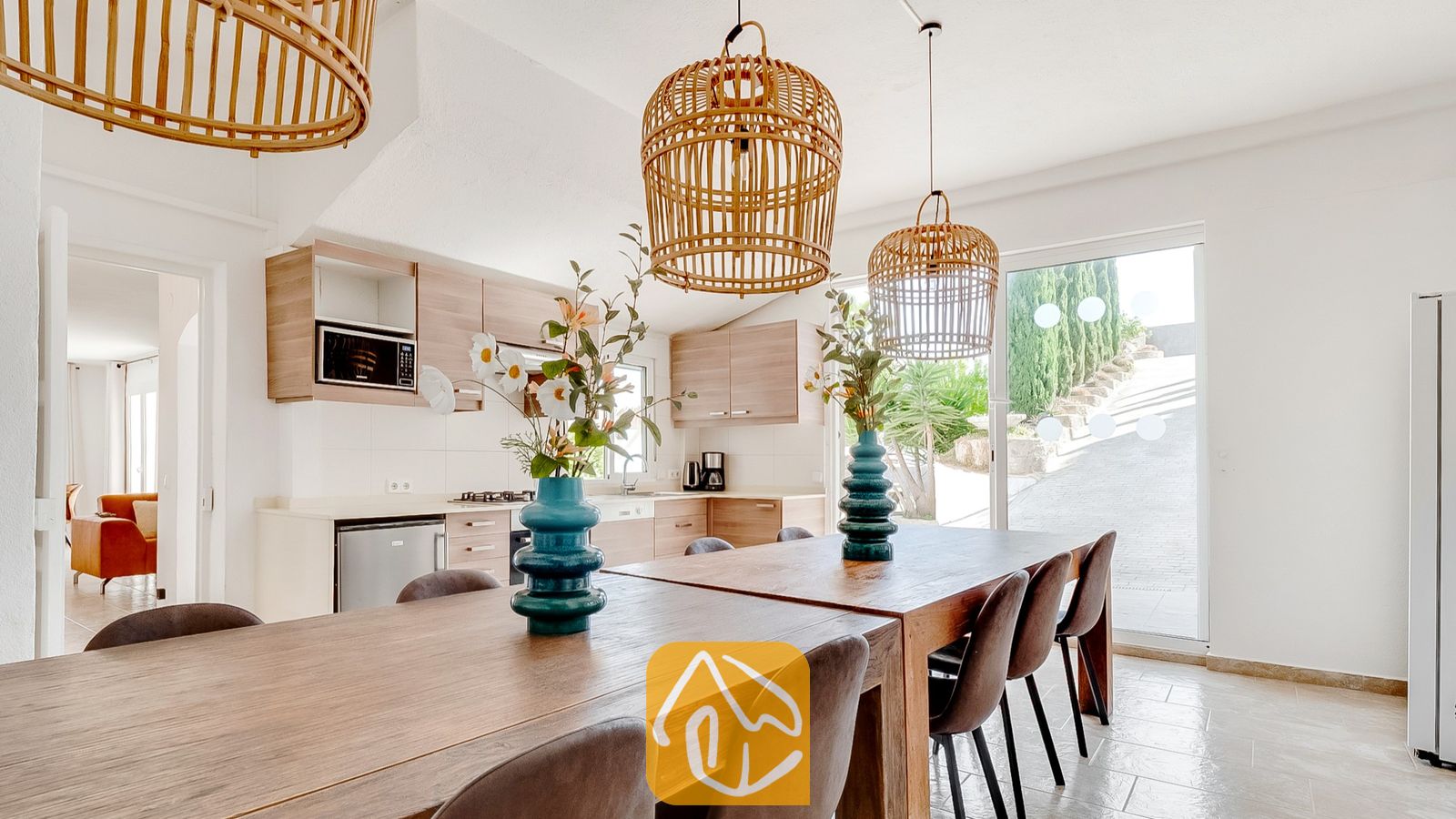 Vakantievilla's Costa Brava Spanje - Villa Tropicana - Houten eettafel met donkere stoelen en turquoise vazen, onder geweven rieten hanglampen in Villa Tropicana, met een keuken zichtbaar op de achtergrond.