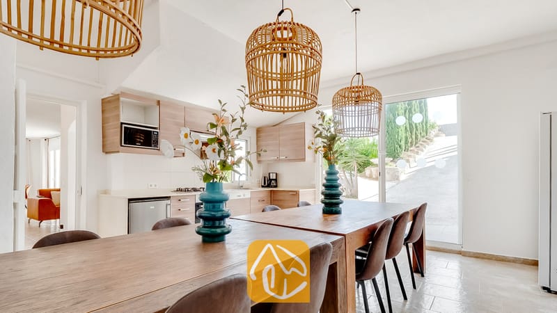 Vakantievilla's Costa Brava Spanje - Villa Tropicana - Houten eettafel met donkere stoelen en turquoise vazen, onder geweven rieten hanglampen in Villa Tropicana, met een keuken zichtbaar op de achtergrond.