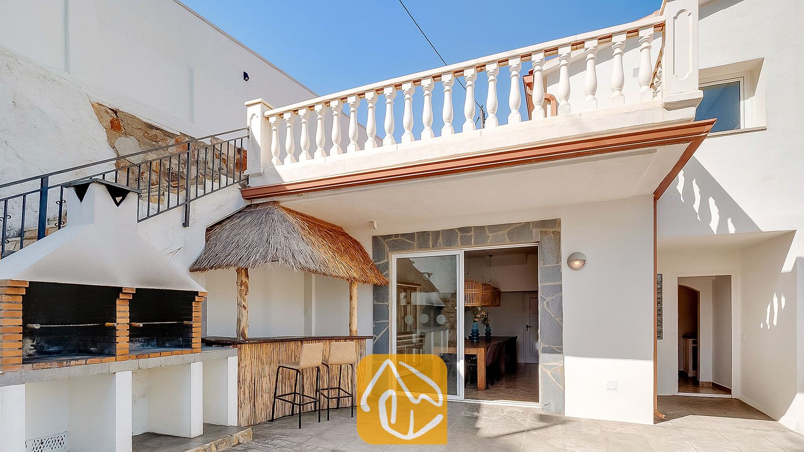 Vakantievilla's Costa Brava Spanje - Villa Tropicana - Buitenterras van Villa Tropicana met een ingebouwde stenen barbecue, een tiki-bar met rieten dak en twee krukken, en een wit balkonhek.