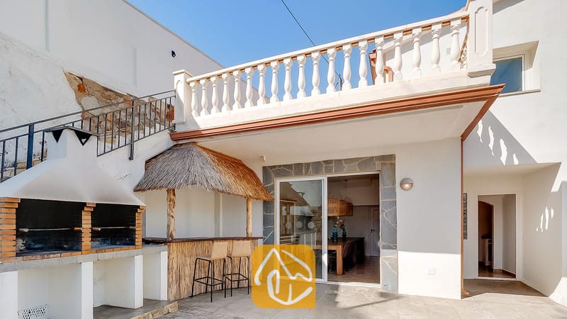Vakantievilla's Costa Brava Spanje - Villa Tropicana - Buitenterras van Villa Tropicana met een ingebouwde stenen barbecue, een tiki-bar met rieten dak en twee krukken, en een wit balkonhek.