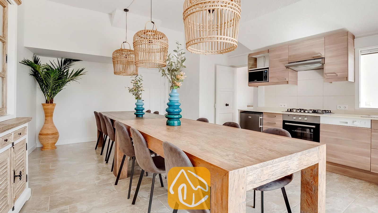 Vakantievilla's Costa Brava Spanje - Villa Tropicana - Villa Tropicana met open keuken en eethoek, voorzien van een lange houten eettafel, grijs gestoffeerde stoelen, rieten hanglampen en licht houten kasten.
