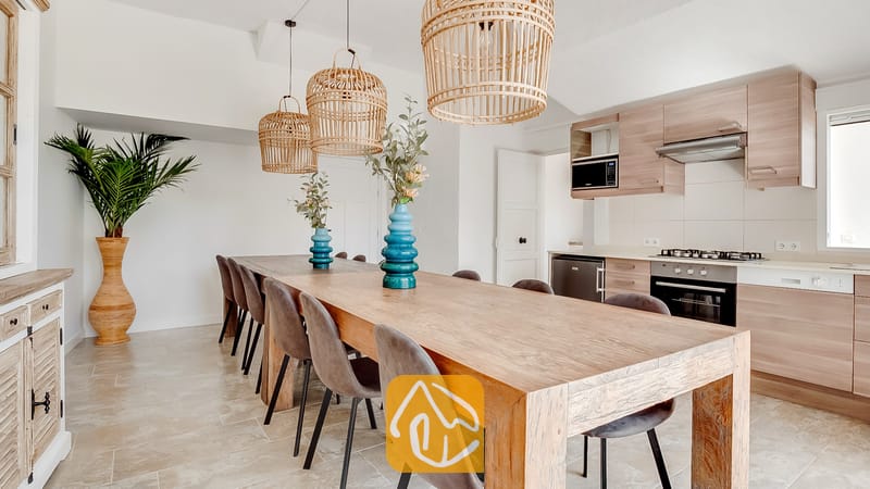 Vakantievilla's Costa Brava Spanje - Villa Tropicana - Villa Tropicana met open keuken en eethoek, voorzien van een lange houten eettafel, grijs gestoffeerde stoelen, rieten hanglampen en licht houten kasten.