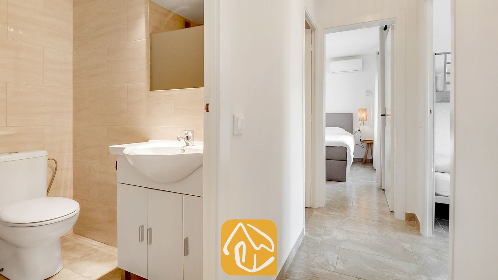 Vakantievilla's Costa Brava Spanje - Villa Tropicana - Badkamer met een wit toilet en wastafelmeubel, die uitkomt op een gang met uitzicht op twee slaapkamers.