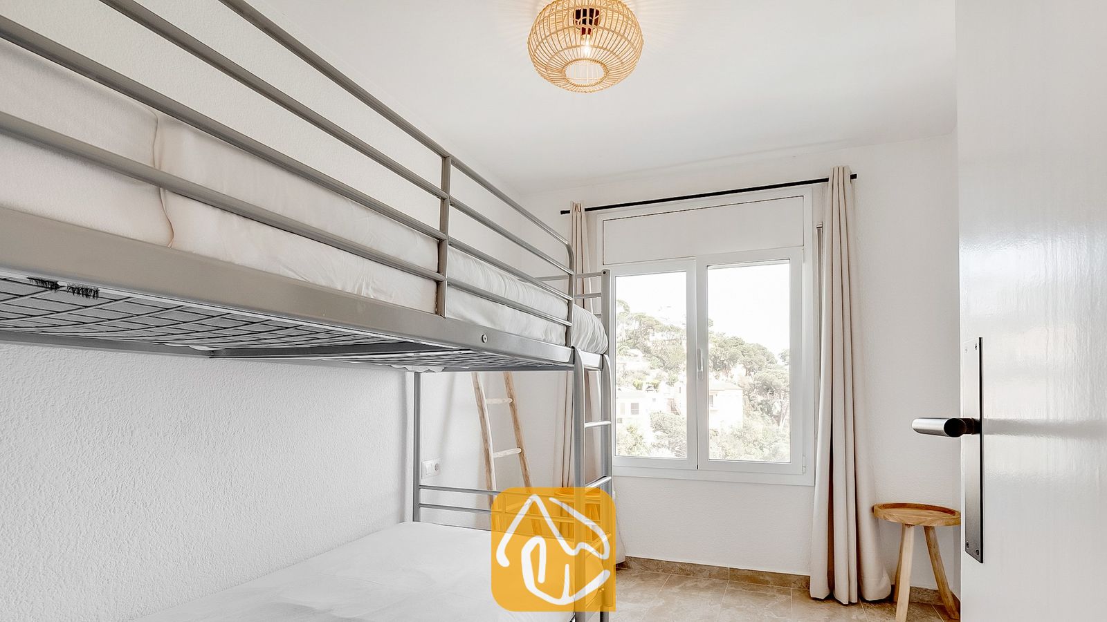 Vakantievilla's Costa Brava Spanje - Villa Tropicana - Slaapkamer met grijs metalen stapelbed, witte muren en een raam met lichte gordijnen dat uitkijkt op bomen.