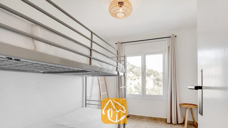 Vakantievilla's Costa Brava Spanje - Villa Tropicana - Slaapkamer met grijs metalen stapelbed, witte muren en een raam met lichte gordijnen dat uitkijkt op bomen.
