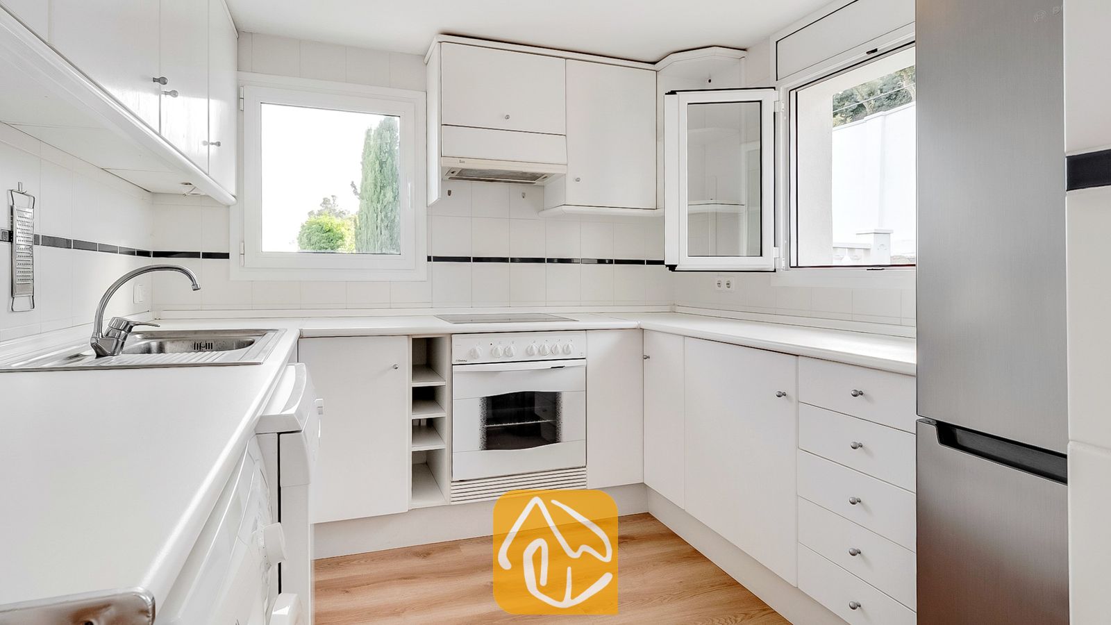 Vakantievilla's Costa Brava Spanje - Villa Tropicana - Licht en fris witte keuken, uitgerust met witte kasten, een inbouwoven met kookplaat, een roestvrijstalen koelkast en twee ramen met uitzicht op de bomen.
