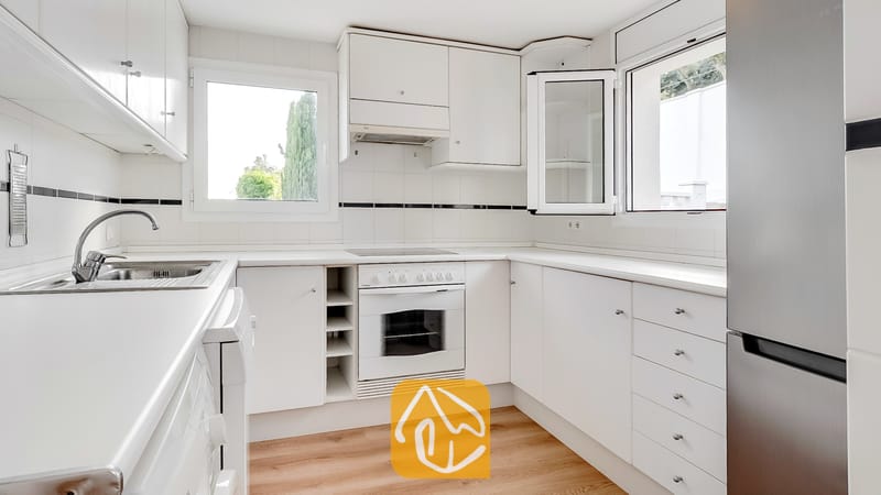 Vakantievilla's Costa Brava Spanje - Villa Tropicana - Licht en fris witte keuken, uitgerust met witte kasten, een inbouwoven met kookplaat, een roestvrijstalen koelkast en twee ramen met uitzicht op de bomen.
