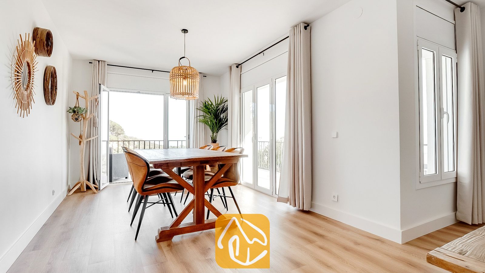 Vakantievilla's Costa Brava Spanje - Villa Tropicana - Eethoek in Villa Tropicana met een houten tafel, bruine stoelen en geweven hanglampen tegen een achtergrond van witte muren en glazen deuren.