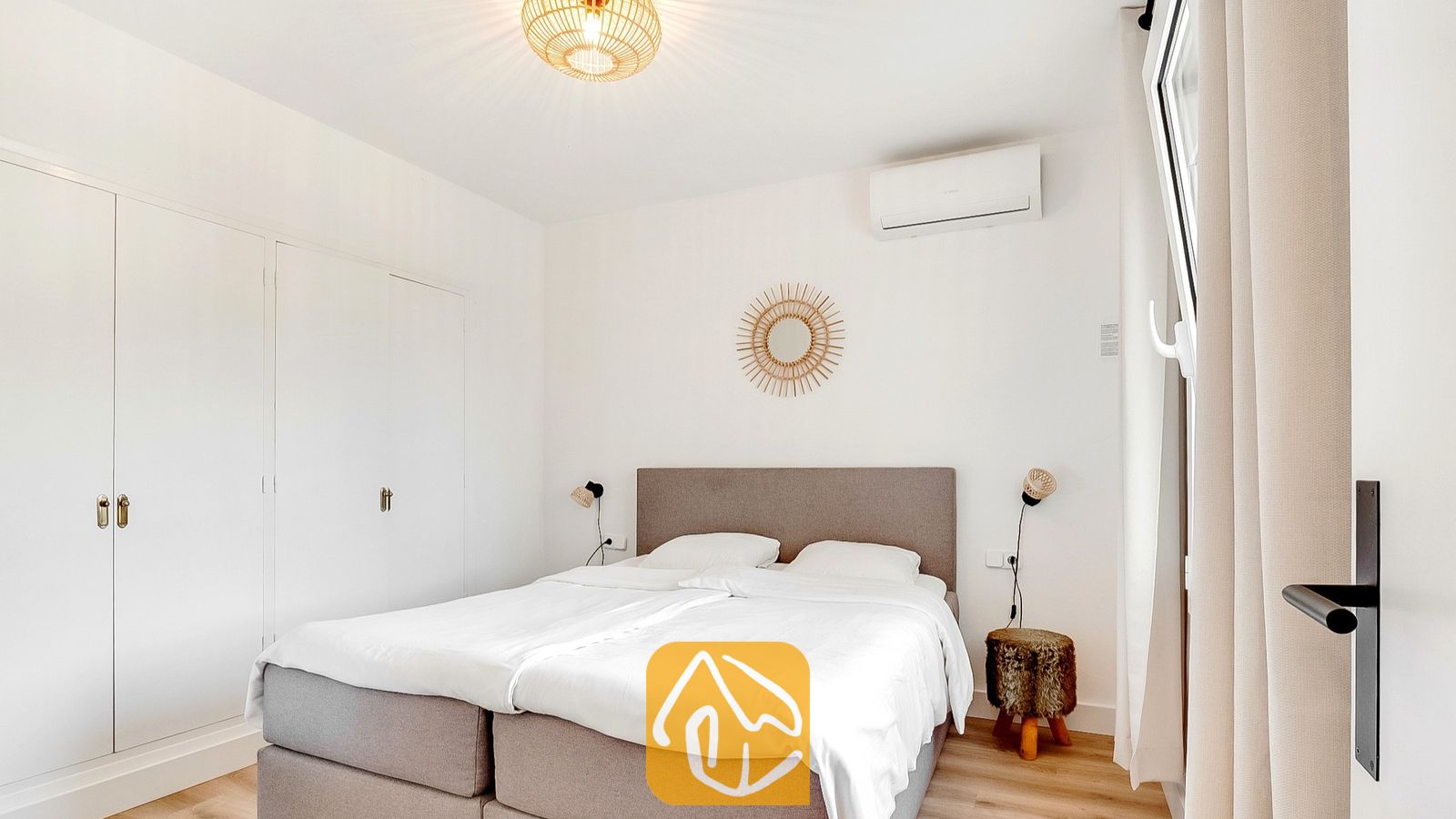 Vakantievilla's Costa Brava Spanje - Villa Tropicana - Slaapkamer met een grijs gestoffeerd tweepersoonsbed, witt linnen en een ronde rotan spiegel in de vorm van een zon op de witte wand.