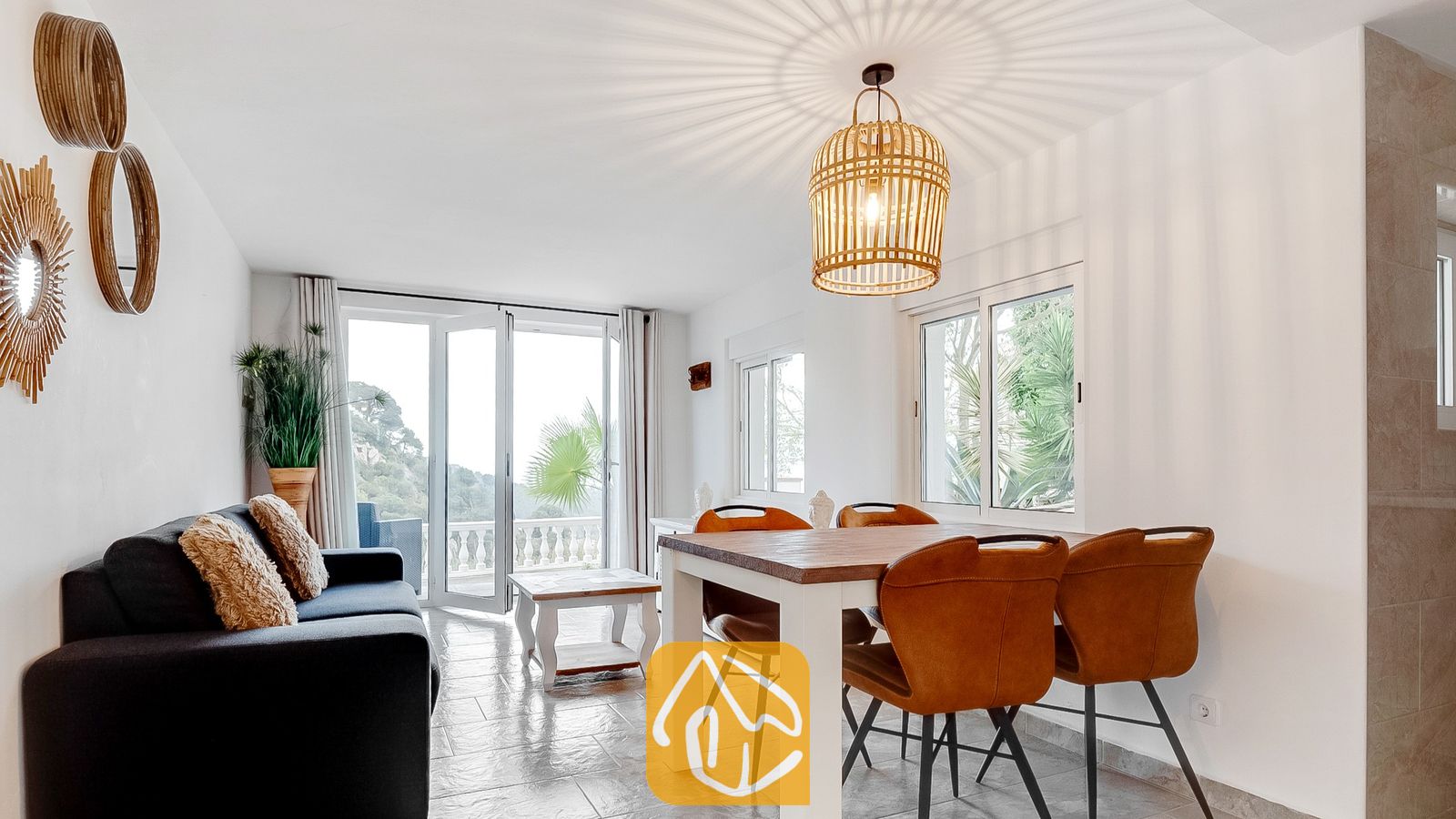 Vakantievilla's Costa Brava Spanje - Villa Tropicana - Woon- en eetkamer van Villa Tropicana met een houten eettafel, bruine lederen stoelen en een donkere bank die uitkijkt op de glazen deuren naar het balkon.