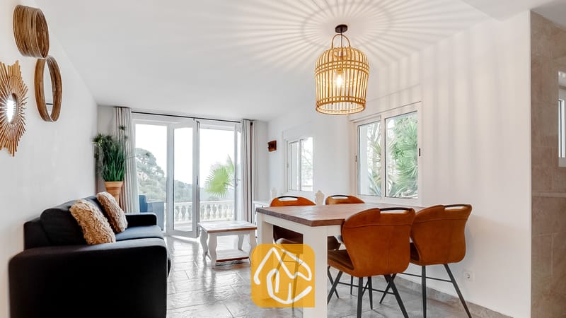 Vakantievilla's Costa Brava Spanje - Villa Tropicana - Woon- en eetkamer van Villa Tropicana met een houten eettafel, bruine lederen stoelen en een donkere bank die uitkijkt op de glazen deuren naar het balkon.