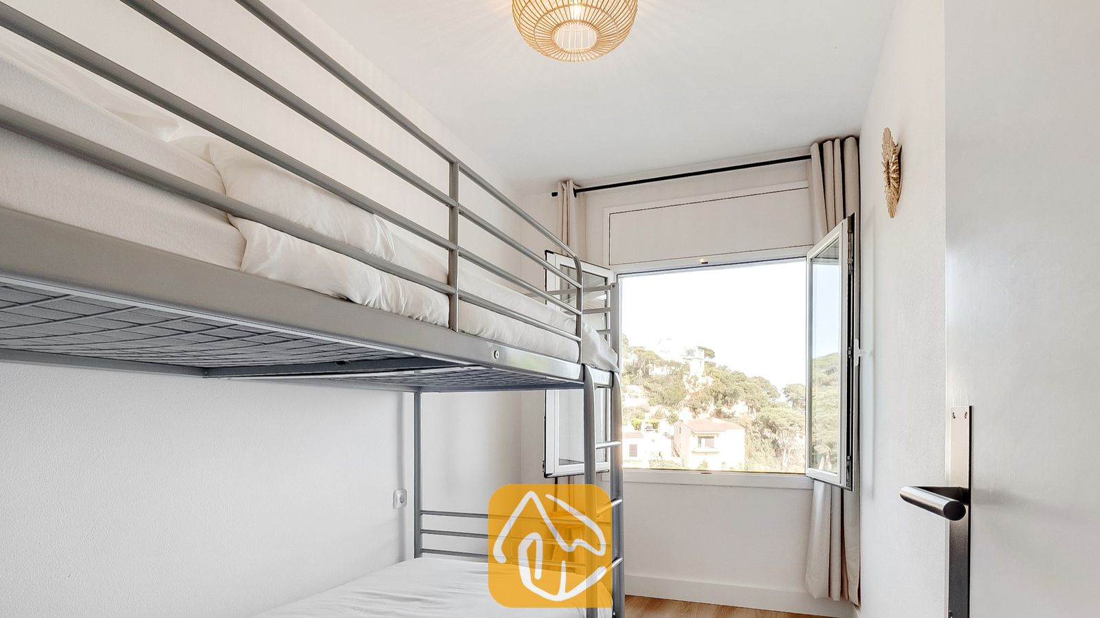 Vakantievilla's Costa Brava Spanje - Villa Tropicana - Slaapkamer met metalen stapelbedden opgemaakt met wit linnengoed, een geweven plafondlamp en een open raam met uitzicht op groen buiten.
