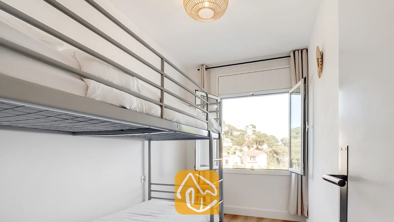 Vakantievilla's Costa Brava Spanje - Villa Tropicana - Slaapkamer met metalen stapelbedden opgemaakt met wit linnengoed, een geweven plafondlamp en een open raam met uitzicht op groen buiten.