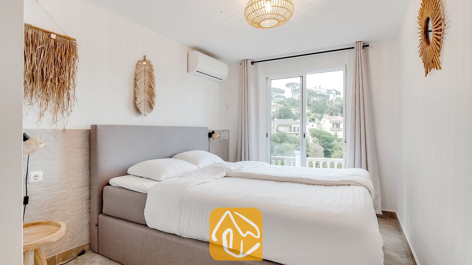 Vakantievilla's Costa Brava Spanje - Villa Tropicana - Slaapkamer met een grijs gestoffeerd bed, wit beddengoed en glazen schuifdeuren die uitkomen op een uitzicht op een groene heuvel.