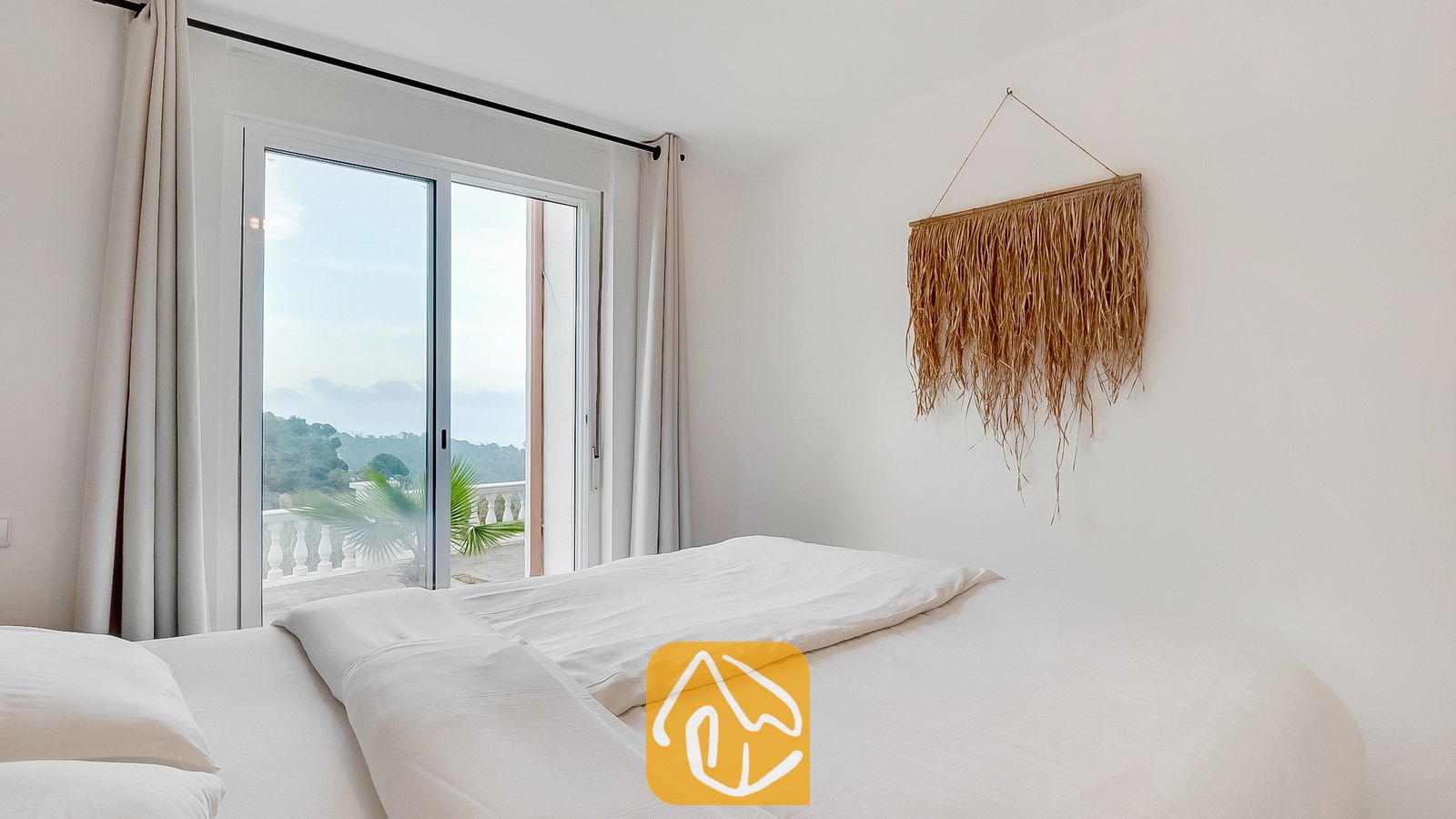 Vakantievilla's Costa Brava Spanje - Villa Tropicana - Slaapkamer met wit beddengoed, een geweven wandkleed van stro en glazen schuifpui die uitkomt op een terras met groen uitzicht.