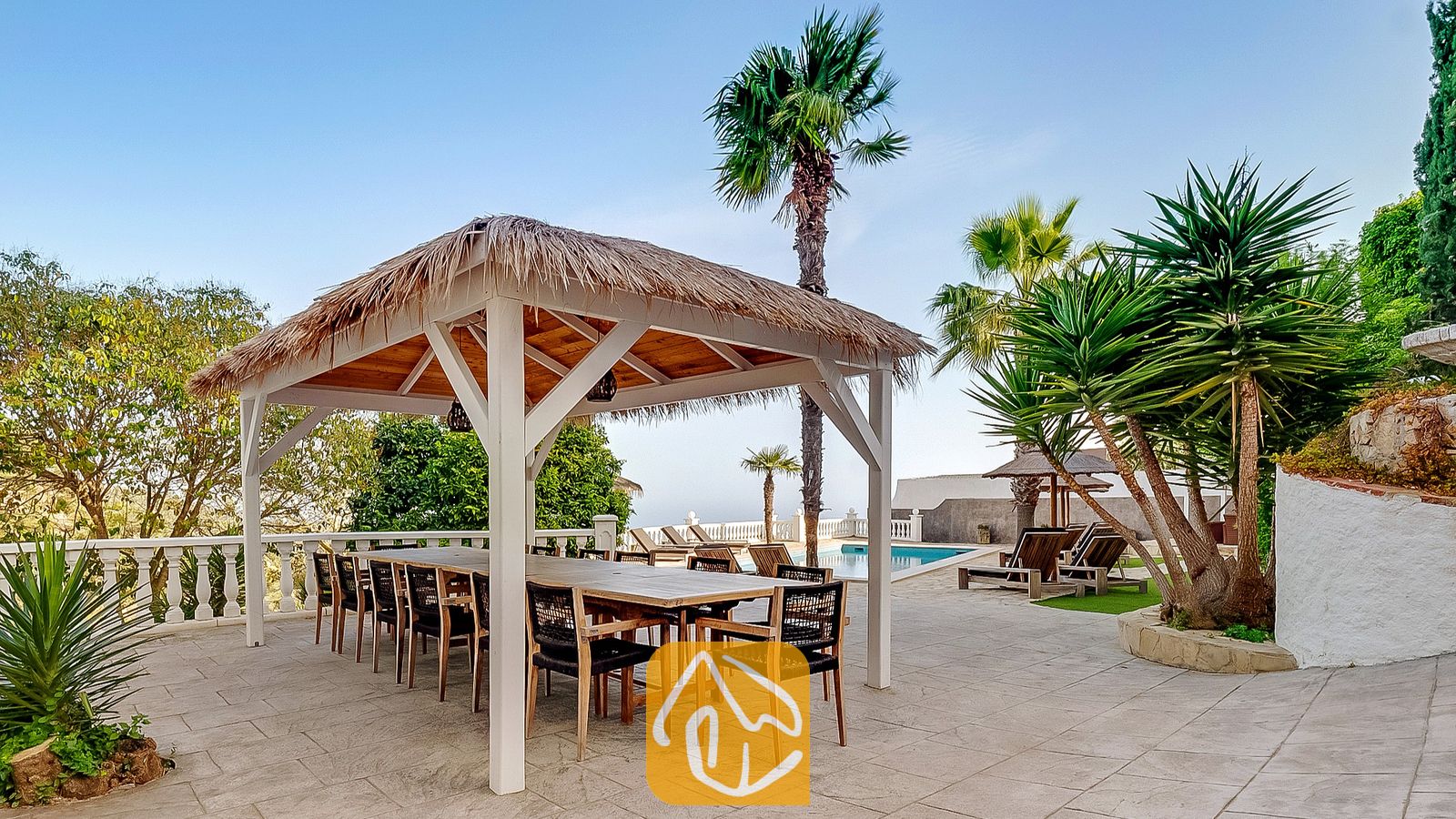 Vakantievilla's Costa Brava Spanje - Villa Tropicana - Overdekte eethoek in een pergola met een rieten dak en een grote houten tafel bij Villa Tropicana, gelegen op een betegeld terras naast palmbomen en een zwembad.
