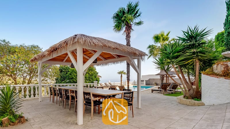 Vakantievilla's Costa Brava Spanje - Villa Tropicana - Overdekte eethoek in een pergola met een rieten dak en een grote houten tafel bij Villa Tropicana, gelegen op een betegeld terras naast palmbomen en een zwembad.