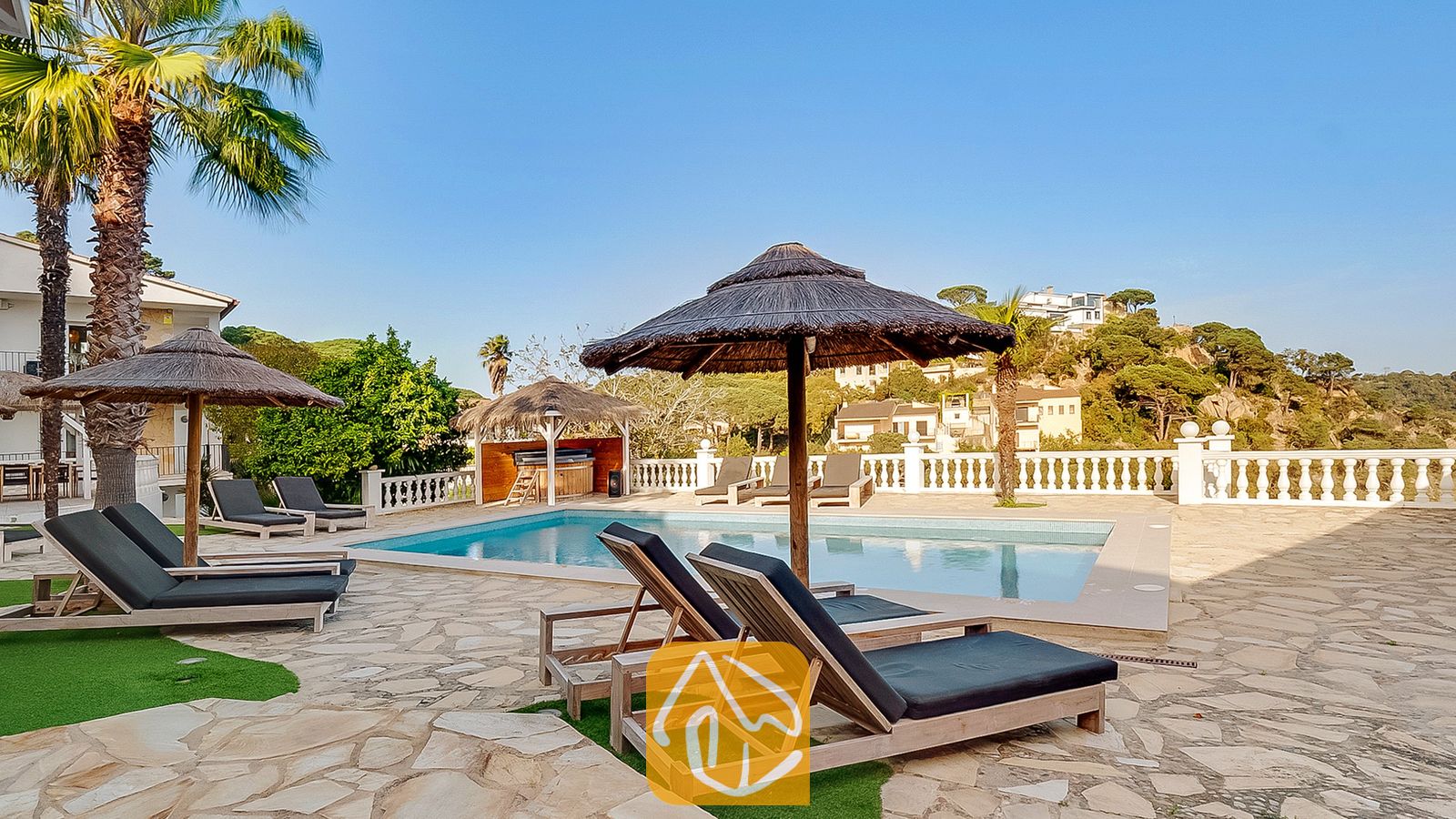 Vakantievilla's Costa Brava Spanje - Villa Tropicana - Zonnig zwembadterras bij Villa Tropicana met houten ligstoelen en donkere kussens, rieten parasols en een zwembad tegen de achtergrond van een heuvel.