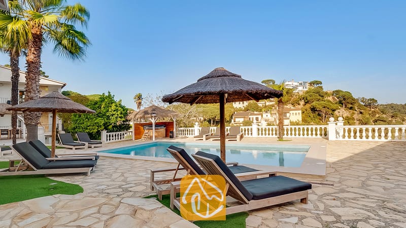 Vakantievilla's Costa Brava Spanje - Villa Tropicana - Zonnig zwembadterras bij Villa Tropicana met houten ligstoelen en donkere kussens, rieten parasols en een zwembad tegen de achtergrond van een heuvel.