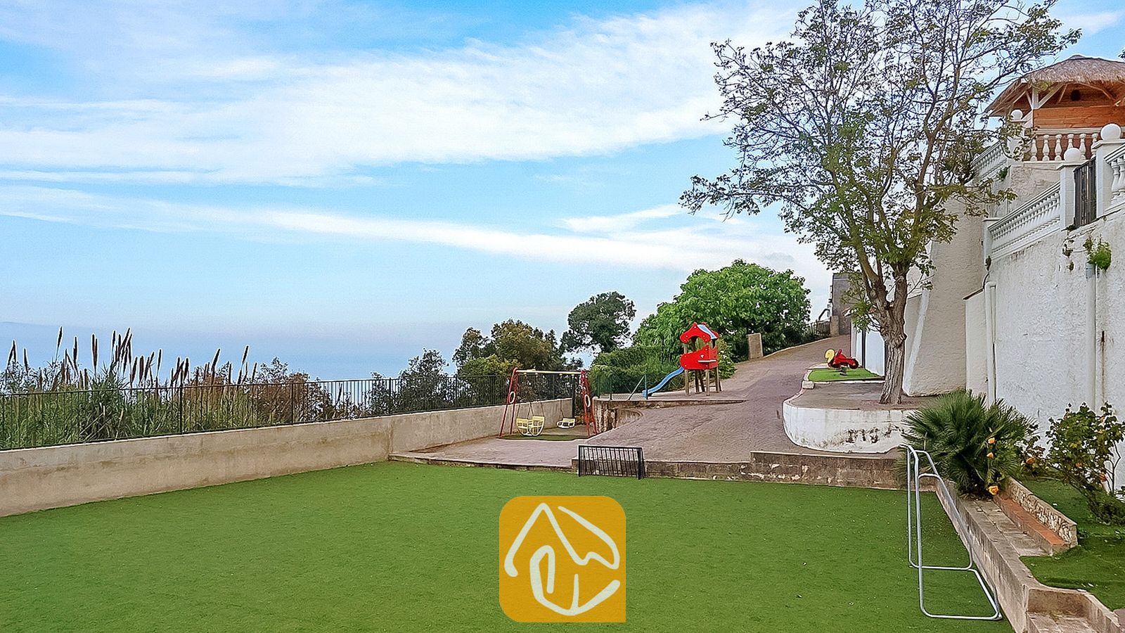 Vakantievilla's Costa Brava Spanje - Villa Tropicana - Buiten speelgedeelte bij Villa Tropicana met een rode schommelset, glijbaan en een groen kunstgrasgazon naast een witte buitenmuur.