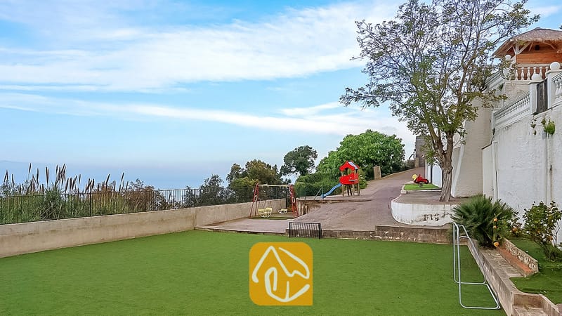 Vakantievilla's Costa Brava Spanje - Villa Tropicana - Buiten speelgedeelte bij Villa Tropicana met een rode schommelset, glijbaan en een groen kunstgrasgazon naast een witte buitenmuur.