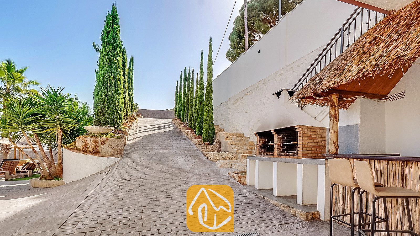 Vakantievilla's Costa Brava Spanje - Villa Tropicana - De op bepaalde oprit van Villa Tropicana is omzoomd door hoge cipressen en beschikt over een stenen barbecue in de buitenlucht en een bar met rieten dak en hoge krukken.