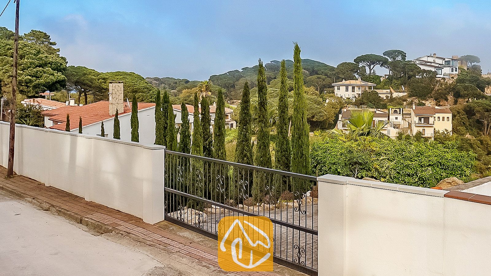 Vakantievilla's Costa Brava Spanje - Villa Tropicana - Buitenaanzicht van Villa Tropicana met een witte tuinmuur, een zwarte metalen oprijlaanpoort en hoge cipressen die uitkijken over een heuvelachtig woonlandschap.