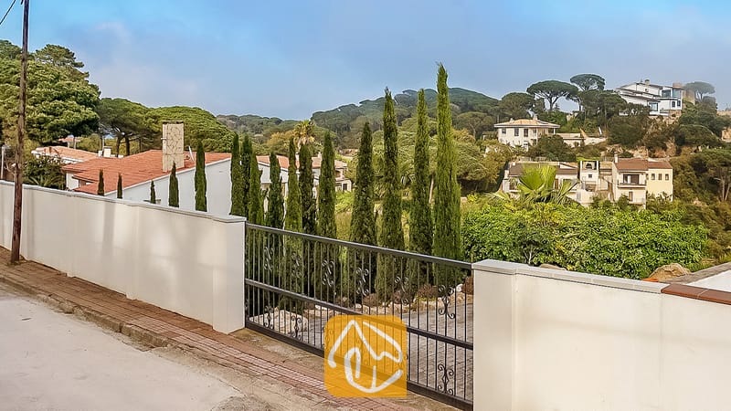 Vakantievilla's Costa Brava Spanje - Villa Tropicana - Buitenaanzicht van Villa Tropicana met een witte tuinmuur, een zwarte metalen oprijlaanpoort en hoge cipressen die uitkijken over een heuvelachtig woonlandschap.