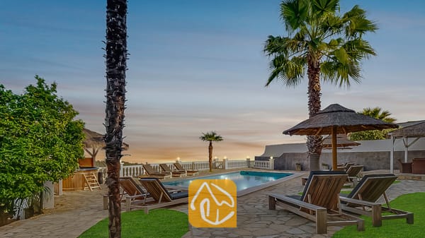 Vakantievilla's Costa Brava Spanje - Villa Tropicana - Privé buitenzwembad bij Villa Tropicana, omringd door houten ligstoelen, palmbomen en rieten parasols onder een avondhemel.