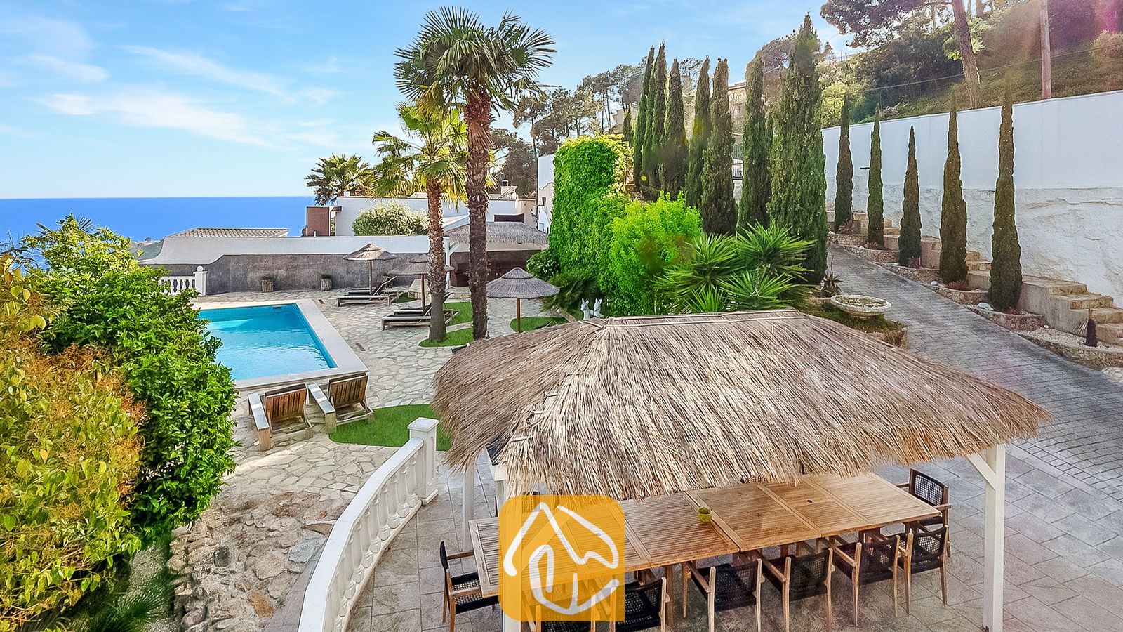 Vakantievilla's Costa Brava Spanje - Villa Tropicana - Buitenkant van Villa Tropicana met een rietgedekte eetpergola, stenen terras, zwembad en uitzicht op zee onder een blauwe hemel.