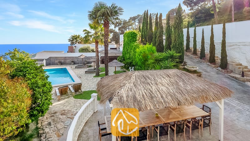 Vakantievilla's Costa Brava Spanje - Villa Tropicana - Buitenkant van Villa Tropicana met een rietgedekte eetpergola, stenen terras, zwembad en uitzicht op zee onder een blauwe hemel.