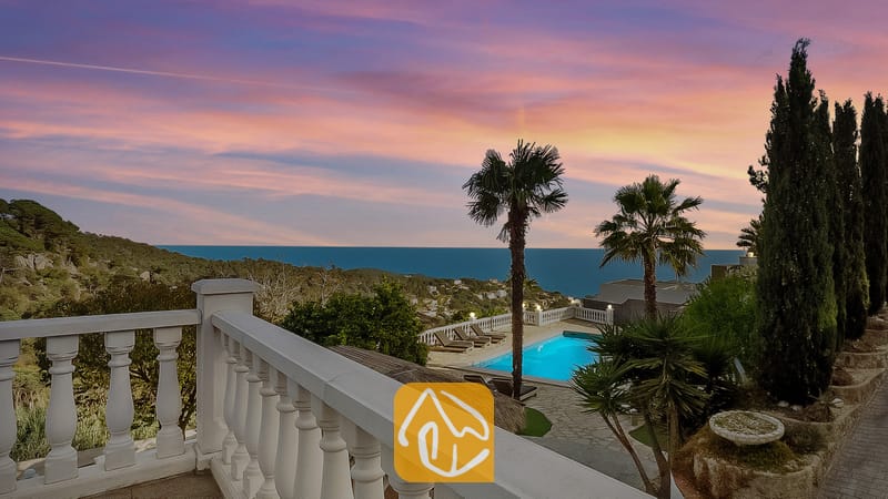 Vakantievilla's Costa Brava Spanje - Villa Tropicana - Terras met uitzicht op Villa Tropicana, voorzien van een eigen zwembad, palmbomen en zeezicht onder een roze en blauwe zonsondergang.