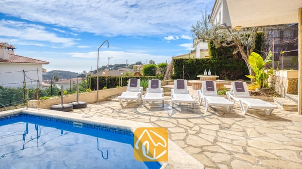 Vakantievilla's Costa Brava Spanje - Villa Janet - Zonnig stenen terras bij Villa Janet met vijf witte ligstoelen naast een blauw betegeld zwembad en uitzicht op een stad in de verte onder een bewolkte blauwe hemel.