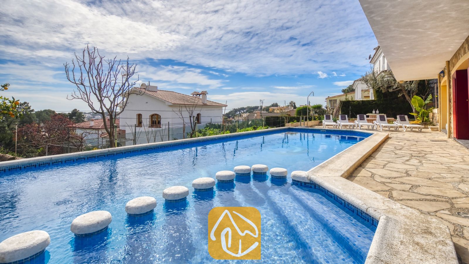 Vakantievilla's Costa Brava Spanje - Villa Janet - Rechthoekig zwembad met stapstenen en een blauw tegelinterieur bij Villa Janet in Spanje, voorzien van een stenen terras en uitzicht op de omliggende buurt onder een bewolkte blauwe hemel.