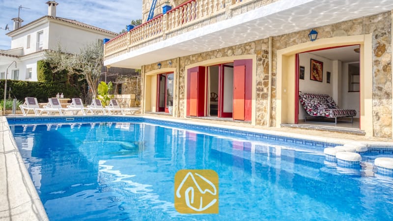 Vakantievilla's Costa Brava Spanje - Villa Janet - Buitenzwembad voor de stenen gevel van Villa Janet met rode deuren, een gemeubileerd interieur dat zichtbaar is door de open deuropening, en ligstoelen op het terras aan het zwembad.