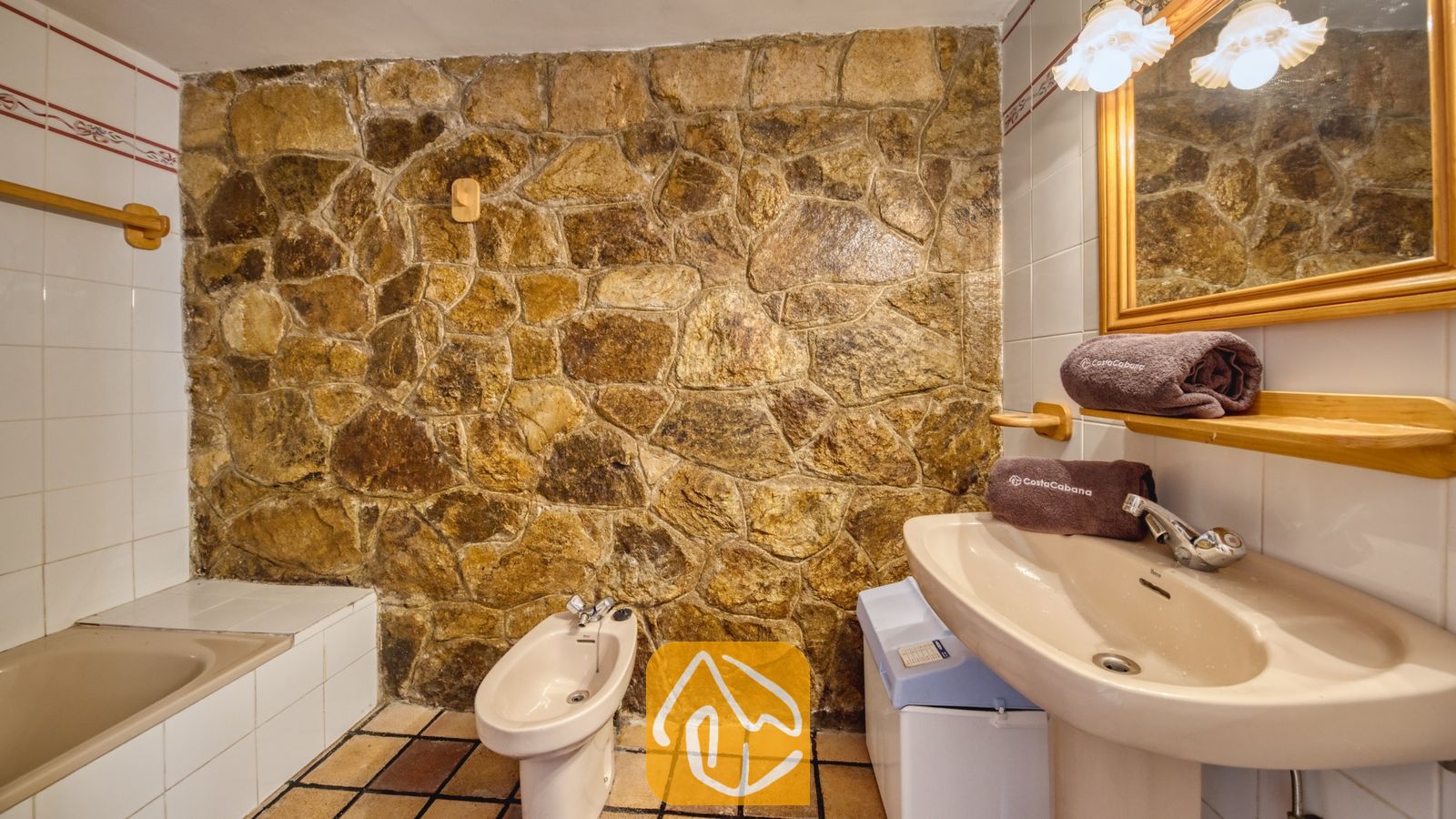 Vakantievilla's Costa Brava Spanje - Villa Janet - Badkamer met een rustieke accentmuur van natuursteen, een beige wastafel op zuil, een bidet en een houten spiegel.