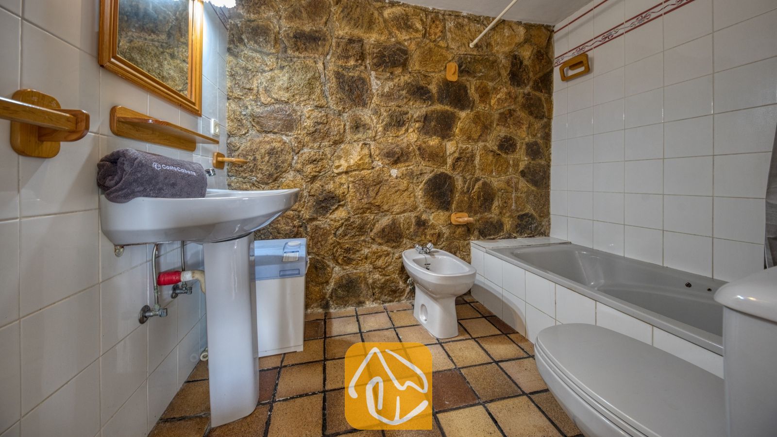 Vakantievilla's Costa Brava Spanje - Villa Janet - Badkamer voorzien van een rustieke accentmuur met blootliggend natuursteen, een witte wastafel op zuil met een houten spiegel, en een tegelvloer.